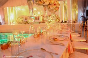 event wedding stage design, signage, chandelier, led dance floor, gold back chameleon chairs, curved VIP table with centerpieces, candles, charger plates, menu card, napkins, drink glassware, event decorator in Nigeria