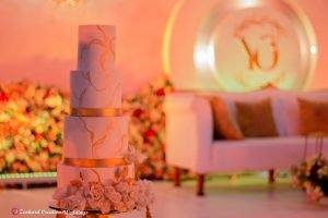 white and gold three tier wedding cake with white rose petals with gold trimmings, white couch with brown pillows, wall washers light and flower arrangement, event planner Eventigo,