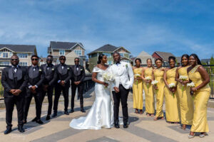 Nigerian white wedding in Canada, white wedding dress gown, white suit on groom with black pant, buttonnaire, white floral bouquet with memorabilia picture of mum, elegant smile, bridal make up, wrist watch, with groomsmen and bridesmaids