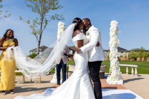 Debola and Okiki white wedding in Canada, white wedding dress gown, white suit on groom with black pant, buttonnaire, white floral bouquet with memorabilia picture of mum, elegant smile, bridal make up, wrist watch, wedding first kiss
