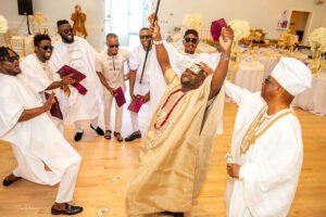 Nigerian couple in Canada traditional wedding, white agbada, champagne gold silk aso oke, walking stick, fila, coral and ivory bead iyun, groom dancing with friends