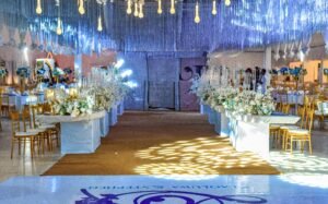 Nigerian couple white wedding reception with monogram dance floor, ceiling installation, VIP high table, chivalry chairs, floral arrangement, 3 in 1 event center hall decoration