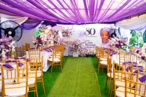 purple and white birthday design for 80th year old grandma with chiavari chairs, green astroturf, flowers, purple table scape, table setting, gold rim plates, purple floral arrangement on tables